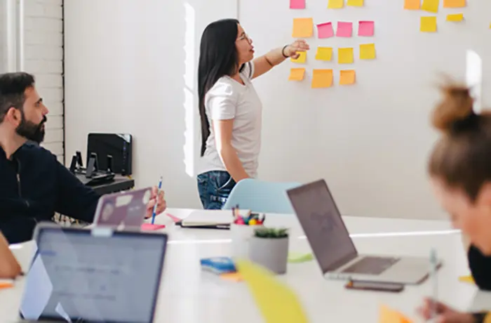 A team of people meeting around a table, with someone standing and pointing at a board with sticky notes on it.