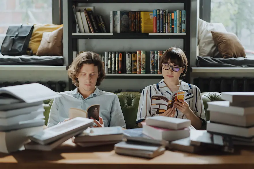 Man and woman sitting next to eachother reading, surrounded by many books